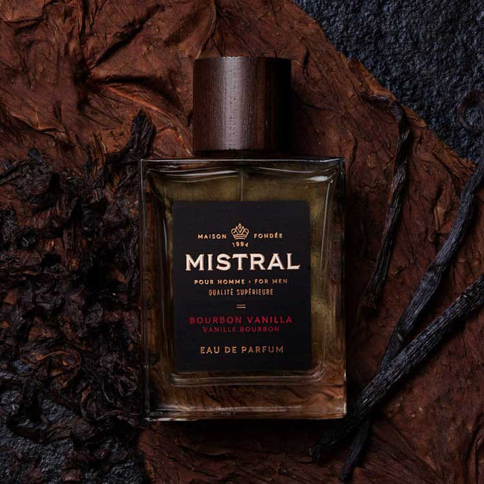 Mistral Bourbon Vanilla cologne in a glass bottle with a dark wood cap, surrounded by tobacco leaves and vanilla beans.