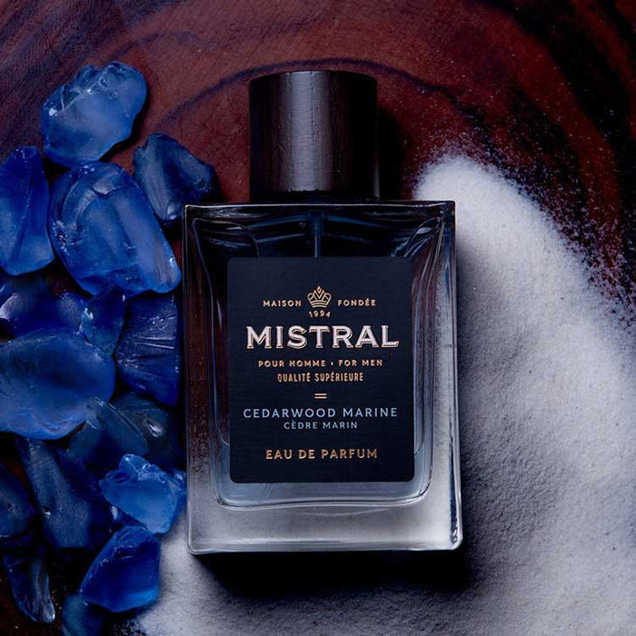 Mistral Cedarwood Marine cologne bottle with a dark cap, surrounded by blue sea glass and white sand on a wood surface.