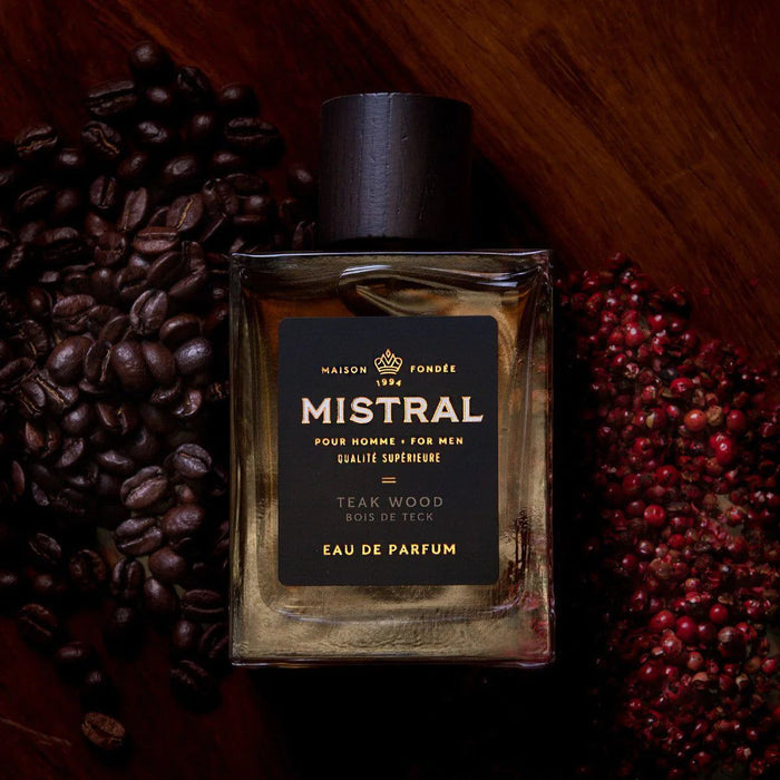 Mistral Teak Wood Eau de Parfum in a glass bottle with a dark cap, surrounded by coffee beans and red peppercorns.