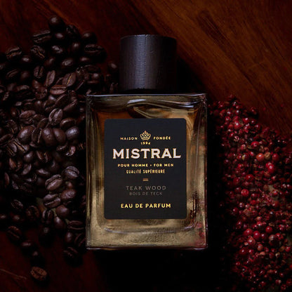 Mistral Teak Wood Eau de Parfum in a glass bottle with a dark cap, surrounded by coffee beans and red peppercorns.