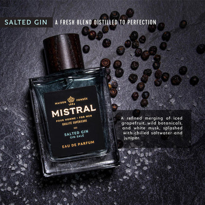 Mistral Salted Gin cologne bottle with a dark wood cap, surrounded by juniper berries and sea salt on a slate surface.